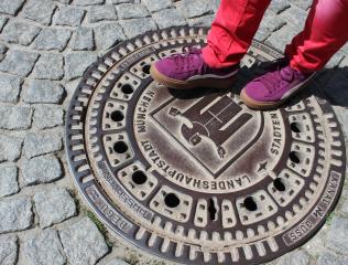 Füße in Turnschuhen auf einem Kanaldeckel mit Münchner Stadtwappen